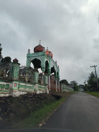 The image depicts the facade of a mosque's entrance structure in Aluepapeun, Indonesia. The structure is painted in vibrant green and white with ornate decorative elements. It features multiple arches and is topped with copper-colored domes, one of which has a crescent moon finial. A metal fence, also painted green and white, runs along the front of the mosque's property.
To the right of the mosque, a paved road stretches into the distance. Power lines and utility poles are visible along the side of the road, with lush green trees and vegetation flanking both sides. The sky is overcast with heavy gray clouds, suggesting a cloudy or rainy day. A dark-colored car is visible on the road in the distance, moving away from the camera. The foreground shows a grassy embankment with some scattered stones and a small ditch. The overall scene is one of a rural or semi-rural setting, with the mosque being a prominent architectural feature.