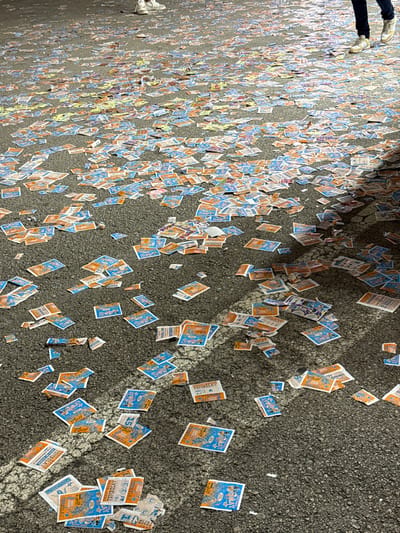 The image displays a dark asphalt ground in Polígon Industrial De Son Castello, Spain, extensively covered with scattered, small, rectangular paper tickets. These tickets are predominantly orange, blue, and yellow-white, many bearing the text "TOMBOLAY" and "PUJA," along with numerical markings. The lower legs and feet of two individuals are visible at the top of the frame; one on the left wears white sneakers and light-colored pants, while another on the right wears dark pants and light-colored sneakers, casting a shadow onto the littered surface. The scene suggests the aftermath of an event involving a widespread distribution and discarding of raffle or lottery tickets.