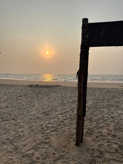 The image depicts a beach scene at sunset in Bhagatwadi, India. The horizon line is visible across the middle of the frame, separating a hazy, light orange sky from the sea. The sun, a bright yellow-orange disc, is low on the horizon, casting a shimmering reflection path on the water. Several small boats are present in the distance on the sea. The foreground features light brown sand with numerous footprints and subtle undulations. In the right foreground, a rustic wooden pole stands vertically, partially wrapped with a darker material, likely rope or fabric. A section of dark, woven material or netting is attached horizontally to the upper part of this pole, extending out of the frame to the right. A small, low sand mound is visible on the beach in the mid-ground. No individuals are present in the scene.