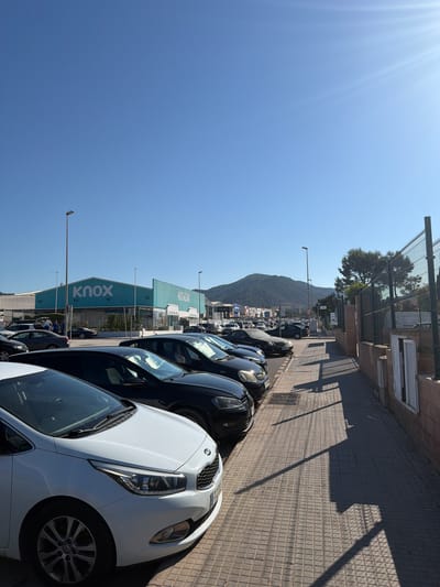 The image shows a row of cars parked on a street in Polígon Industrial De Calvià, Spain. The street is paved with brick tiles, and there are shadows cast by a fence and a building. The cars are parked in a line along the street. In the background, there are several buildings, one of which has the word "KNOX" written on it in large blue letters. There is also a mountain in the distance. The sky is clear and blue, indicating a sunny day. The sun is shining brightly, creating lens flare in the upper left corner of the image. The time of day appears to be daytime, likely late morning or early afternoon, given the length and angle of the shadows. No people are clearly visible in the image. The scene suggests a commercial or industrial area.