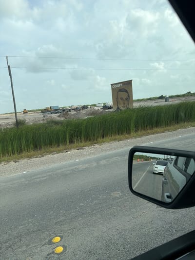 The image captures a scene from a car traveling on a road in Brownsville, Texas, USA. In the foreground, a side-view mirror reflects a white Tesla and other vehicles on the road. Beyond the road, a field of tall, green grass lines the side of the road. In the background, a large mural of a man's face with the words "OCCUPY MARS" above it is visible on a tall structure. The landscape appears to be a construction or industrial site, with heavy machinery, pipes, and piles of dirt scattered across the area. The sky is overcast with clouds.