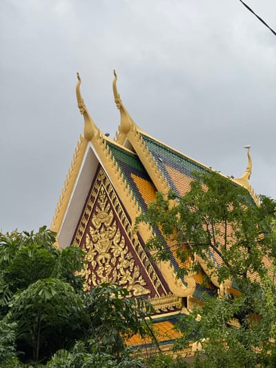 This media file shows a low-angle shot of the ornate roof of a building, likely a temple, in Cambodia. The roof is adorned with intricate golden carvings against a dark red background, with colorful tiles in shades of green, yellow, and blue. The building's eaves are decorated with a repetitive serrated pattern, and the roof curves upwards to pointed finials. Lush green foliage from surrounding trees partially obscures the building, suggesting an outdoor setting. The sky above is overcast, indicating a cloudy or possibly rainy day. A thin black line, potentially a wire, is visible in the upper left corner of the frame. No people or text are visible in the image. The style of the architecture and the foliage point to a tropical or subtropical location, consistent with the provided context of Khan Daun Penh, Cambodia.