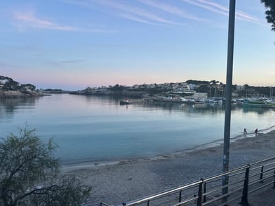 The image depicts a coastal scene in Porto Cristo, Spain. The foreground includes a stainless steel railing and a partially visible tree on the left side. A narrow beach composed of sand and pebbles leads to calm water, which fills the central portion of the image. In the distance, a small town is visible along the coastline, featuring white buildings and some vegetation. A small pier with a red lighthouse stands near the town. Several boats and yachts are docked in the harbor area. Two people are seen walking along the beach near a light pole. The sky displays hues of blue and pink, suggesting either early morning or late evening.