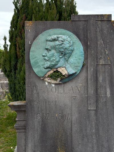 This image depicts the gravestone of J.B. Yollant, an architect, located in Auvers-sur-Oise, France. The gravestone is made of grey stone and includes a bronze medallion featuring a profile of Yollant. The medallion is partially broken, with small plants growing in the damaged section. Text carved into the stone reads "J.B. YOLLANT ARCHITECTE" and "1852-1894". The signature "Fagel 1875" is inscribed on the medallion. A dense evergreen tree or shrub is visible in the background to the left of the gravestone. The sky is overcast. Other graves and vegetation are vaguely visible in the background. The orientation of the image is rotated 90 degrees counter-clockwise.