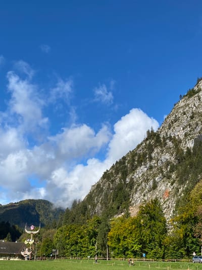 This image depicts a mountainous landscape and a recreational area in Abondance, France. The scene is dominated by a clear blue sky with scattered white cumulus clouds, occupying the upper half of the frame.
On the right, a steep, rocky mountain face rises, characterized by grey and white rock formations and sparsely covered with green coniferous trees. A small patch of reddish-orange rock is visible on the lower right section of this slope. To the left, in the background, a range of forested hills is partially obscured by mist or low clouds.
The midground features a vibrant green grassy field, delineated by white tape or rope markings. A line of mixed deciduous and coniferous trees, exhibiting autumn colors with green and yellow foliage, separates the field from the base of the rocky mountain.
On the far left side of the field, a large, stylized sculpture, known as the Vache Abondance (Abondance Cow), stands prominently. It is a multi-component, metallic structure resembling a bovine figure with large, upward-curving horns, dark circular "eyes," and a central body composed of various abstract shapes. Adjacent to and partially obscured by the sculpture is a low-rise building with a dark roof and light-colored walls.
Several small human figures are sparsely distributed across the green field; one figure appears to be kneeling or bending over, while others are standing or walking. On the far right edge of the image, a silver vehicle is parked next to what appears to be a road, with a blue parking sign visible nearby.
