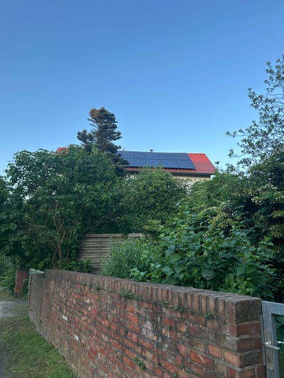 In Schöningen, Germany, a residential building with a red-tiled roof and solar panels is partially visible above lush green foliage. A weathered red brick wall runs along the foreground, with grass and a paved area on its left. A metal gate is situated at the right end of the wall. The sky is clear and blue, with a few faint specks that could be birds or dust.