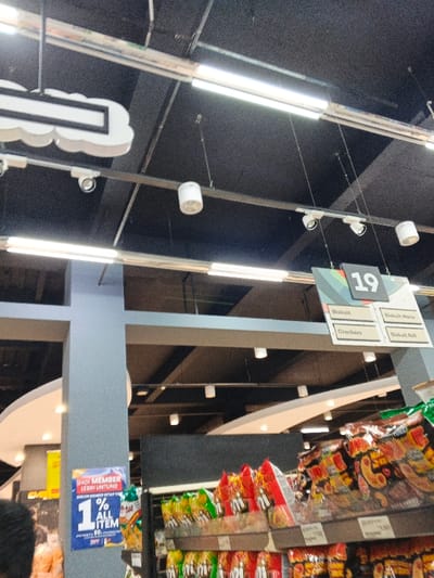 The image captures an indoor scene within what appears to be a supermarket or grocery store, characterized by high ceilings and organized product displays. The dark, industrial-style ceiling is crisscrossed with metal beams and tracks, from which numerous fluorescent tube lights and smaller white spotlights are suspended, illuminating the area. A decorative cloud-shaped light fixture or sign is visible in the upper left.

In the mid-ground, a gray structural pillar stands prominently on the left. Attached to this pillar is a blue sign with white text, reading "JADI MEMBER LEBIH UNTUNG" (Become a member, more profitable) and "1% ALL ITEM," suggesting a membership program offering discounts. To the right, a hanging sign with the number "19" categorizes products below into "Biskuit," "Biskuit Marie," "Crackers," and "Biskuit Roll."

The lower right portion of the image shows several shelves laden with packaged snacks, predominantly chips and possibly instant noodles, in vibrant red and green packaging. Pricing labels are partially visible on the shelves, with one clearly displaying "11.500." A small portion of a person's head is seen in the bottom left corner. The setting implies a commercial retail environment in Aglipay, Philippines, likely during store operating hours. No specific activity or strong emotions are discernible.