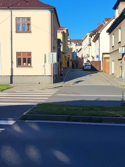 The image shows a street scene in Nový Jičín, Czechia. A cobblestone street leads uphill between buildings. In the foreground, a pedestrian crossing is visible. To the left, a cream-colored building has two windows. The upper window is divided into multiple panes, and the lower window has a red frame and a circular logo inside. A person is standing on the sidewalk in the middle ground. A white car is parked on the street. Signs for parking and the "PRAMOS" company are visible on the right side of the street. The sky is clear and blue.