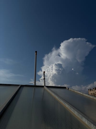 The image shows a view from a rooftop in Ugento, Italy. In the foreground, there is a sloped, translucent roof structure with a metal frame. Two vertical pipes rise from the roof; the taller pipe has a metallic band near its opening, and the shorter pipe has an orange cap and a black band around it. A thin wire runs diagonally across the scene from the left. The sky above is a deep blue, with a large, dramatic cumulonimbus cloud formation dominating the right side of the frame. In the far background, to the right, a stone building with crenellated battlements is partially visible. The scene appears to be captured on a bright, partly cloudy day.