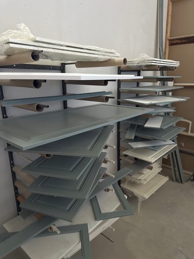 In the city of Kizilyurt, Russia, a workshop is depicted where cabinet doors are being manufactured. The scene is dominated by shelves filled with numerous cabinet doors, many of which are a muted gray-blue color and feature a raised panel design. Some of these doors are stacked horizontally, supported by metal shelving units. Rolled-up paper or protective wrapping materials are visible on some of the shelves, likely used for transport or protection of the finished products. In the background, the environment appears to be a workshop or factory setting, with plain walls and what looks like additional shelving or storage units. Some of the cabinet doors are covered in plastic wrapping, suggesting they are either in the process of being protected or have recently undergone a finishing process. The overall impression is one of an organized workspace dedicated to the production of custom cabinetry.