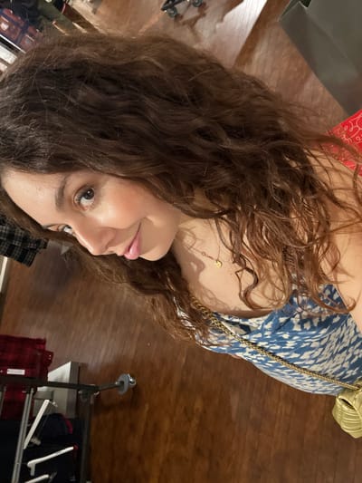 A woman with curly brown hair faces the camera. She wears a blue patterned top with a gold chain strap and matching leaf-shaped bag. A delicate gold necklace adorns her neck. The background features a wooden floor and clothing racks holding red and blue items. The image appears to be taken indoors with warm lighting.