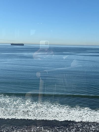 The photo captures a seascape view from a window, revealing the ocean’s surface with slight ripples. A clear blue sky complements the water. A dark structure, possibly a barge, is visible on the horizon. Reflections on the window create an overlay effect. Along the right edge, a textured area of white and gray suggests the shoreline or the sea foam of waves. The overall perspective is elevated, lending a broad view of the horizon and the water's expanse.