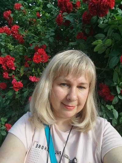 A young woman with blonde hair and blue eyes smiles at the camera. She is wearing a light pink t-shirt with a graphic print on the front. The woman is positioned in front of a bush with bright red roses. The image appears to be taken outdoors, possibly in a garden or park, with the natural light suggesting it was taken during the day. The location is identified as Chernorech'e, Russia, indicating a potentially rural or suburban setting.