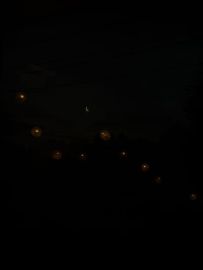 The image depicts a dark night sky over Jāņupe, Latvia, with a thin crescent moon visible in the upper center. A string of illuminated bulbs hangs across the frame, casting a warm, yellow glow against the blackness. The bulbs are spaced at irregular intervals, creating a somewhat whimsical and festive atmosphere. In the background, the silhouette of trees and possibly power lines can be discerned, suggesting an outdoor setting, likely in a rural or suburban area of Jāņupe. The overall mood is one of quiet serenity and enchantment.