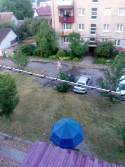 This is a medium-angle, slightly blurred, eye-level shot of an urban scene in Tiachiv, Ukraine. In the foreground, a thick, white rope with black speckled patterns stretches diagonally across the frame, obscuring the view of the area below.
The background reveals a residential building with a light-colored facade and multiple windows. A red balcony juts out from the building, with laundry hanging on a line. Below the building, a paved area or road is visible, with several cars parked. One car, a silver sedan, is parked in the middle of the frame. A person, possibly a woman in a light-colored dress, is walking on the pavement. Green trees and bushes are scattered throughout the scene, adding a touch of nature to the urban environment. In the lower right portion of the frame, a blue, polygonal umbrella lies on its side on a patch of dry grass. In the immediate foreground, the edge of a purple, corrugated rooftop is visible.
The overall impression is of a quiet, ordinary day in a residential area. The lighting suggests it might be overcast or late afternoon. There is no visible text in the image.