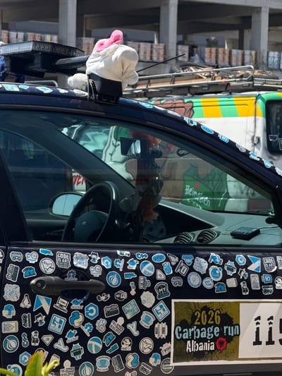 This close-up, vertical shot captures the side of a dark blue vehicle, likely a car, adorned with numerous stickers. The stickers are predominantly blue and white, featuring various logos, text, and icons related to programming and technology, such as "docker," "JS," "HTML," "PHP," and "algorithm." Some stickers express humor or opinions, like "THINGS I HATE" and "CODE IS ART THAT DOES SOMETHING." A larger sticker prominently displays "2026 Carbage run Albania."

The vehicle's window is open, revealing the interior. A black steering wheel is visible, along with an air vent and a side mirror. Hanging from the driver's side mirror is a white stuffed object with a pink bow, resembling a plush toy or decoration.

In the background, slightly out of focus, is a truck with a colorful design featuring yellow, green, and white stripes. The scene appears to be outdoors, under daylight conditions. The setting includes visible building structures with concrete balconies and what looks like stacks of goods or products on the left side of the image. The overall impression is a candid street-side view of a personalized vehicle.