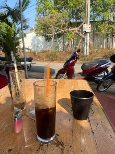 The image depicts an outdoor cafe scene in Đak Đoa, Vietnam, under bright daylight. In the foreground, a weathered wooden table displays three drinking vessels: a clear glass containing dark liquid remnants and a clear straw on the left, a central clear tall glass approximately half-full with a dark beverage topped by a light-colored foam and ice, containing an orange straw, and an empty black fluted plastic cup on the right. A pink and silver lighter is positioned to the left of the central glass, and a white cigarette pack, partially visible, rests beneath it. Beyond the table, a paved road is visible with several parked motorbikes, including two dark red Honda scooters (one partially obscured) and a blue scooter. The background features a white building wall partially obscured by deciduous trees with green foliage and a utility pole with electrical components. Palm-like vegetation is present on the far left. No individuals are visible.