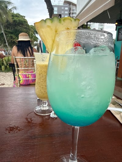 An outdoor table at a restaurant in Jacó, Costa Rica, displays two drinks on a wooden surface. In the foreground, a large wine glass holds a vibrant blue and turquoise cocktail filled with ice and garnished with a slice of pineapple and a red cherry. To the left, a tall glass contains a yellowish-green smoothie or blended drink. In the background, a person wearing a straw hat and a colorful, patterned swimsuit is seated at a chair, facing away from the camera. Palm trees and tropical foliage are visible behind the person, and the sky is overcast. Buildings are faintly discernible in the distance.
