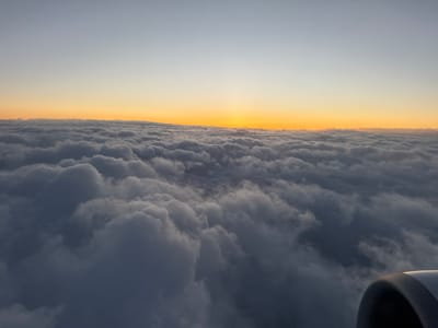 An elevated view from an aircraft shows a dense layer of clouds with the setting sun glowing golden and orange above them. The sky transitions from a light gray to a soft orange near the horizon. The clouds below are a mixture of gray and white, appearing fluffy and textured. A portion of the aircraft's engine is visible in the bottom right corner. The image does not depict Lakatnik, Bulgaria, nor does it show people or actions beyond the flight itself.
