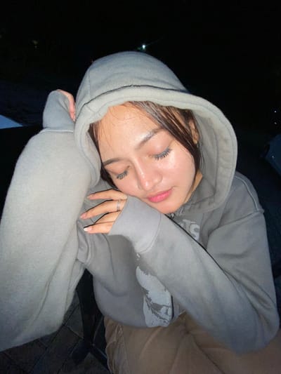 The image shows a young woman wearing a grey hooded sweatshirt and light brown pants. She is leaning her head on her arm, with her eyes closed, appearing to be asleep or resting. A silver ring is visible on her left ring finger. The background is dark and out of focus, suggesting it is nighttime. There is a paved surface with brick patterns visible in the lower portion of the image. The overall scene is a close-up portrait of the woman, with minimal environmental details. The location is not discernible from the image itself; any assertion of it being in Mekar Bakti, Indonesia, is external information.