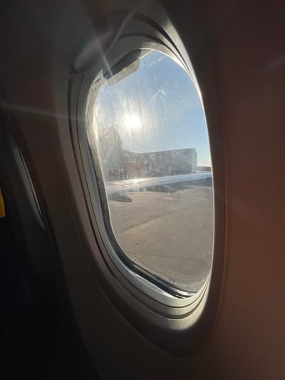 The image depicts a forensic view from an oval-shaped airplane window, looking outward onto an airport apron in Son Santjoan, Spain, consistent with Palma de Mallorca Airport. The scene is illuminated by intense sunlight, positioned in the upper left of the window, creating significant glare and reflections on the dirty window glass, which exhibits numerous smudges and specks. Faint text, possibly "S-0722-30017", is visible on the windowpane.

A portion of the aircraft's wing is visible in the mid-ground, extending horizontally across the lower half of the frame, its metallic surface reflecting the bright sunlight. Beyond the wing, a paved dark gray tarmac or apron surface is visible. In the background, a multi-story airport terminal building or a large structure, possibly connected to a jet bridge, stands under a clear, bright blue sky. The aircraft appears stationary, parked on the apron. The interior cabin elements surrounding the window are visible in warm beige and darker tones, with a hint of a dark seat and yellow detail on the far left. No individuals are discernible within the frame.