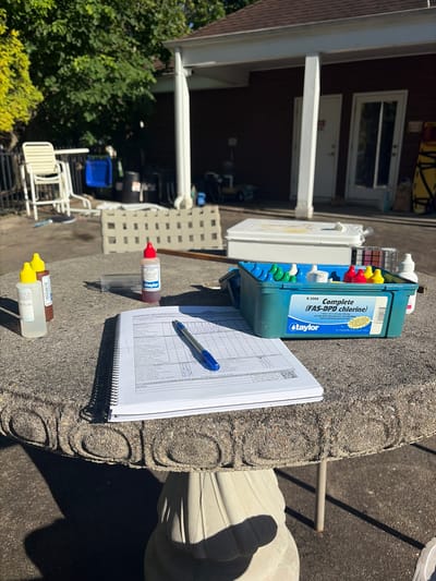 This outdoor scene shows pool maintenance and testing equipment on a textured grey table. The equipment includes a Taylor K-2006 FAS-DPD chlorine test kit in a blue case with various colored caps on reagent bottles, a clear testing vial with a red-capped reagent bottle nearby, and a pair of yellow-capped bottles. A spiral-bound notebook is open next to the equipment, with a blue pen resting on its pages filled with columns and text related to pool testing and maintenance. In the background, there is a building with open windows and white trim. A set of stacked white chairs and a blue plastic tub are visible near a metal fence. Trees and greenery surround the area. The photograph was taken in Edison, United States of America.