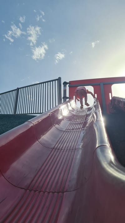 A child in pink attire is at the top of a red metal slide located in an outdoor playground in Kiryat Gat, Israel. The slide is positioned against a blue sky with scattered clouds. A metal fence is visible near the slide. The sun is positioned in the upper left corner of the image, causing a glare on the slide's surface.