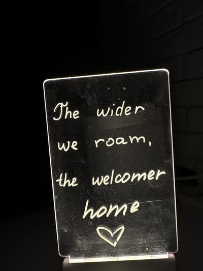 A lit sign with white cursive text is positioned in front of a dark background. The text reads: "The wider we roam, the welcomer home." Below the text, a simple heart outline is drawn. The sign itself appears to be made of a reflective material, possibly acrylic, and has a subtle grid pattern etched into its surface. The lighting creates a glow around the text and the edges of the sign. The scene is devoid of any people or specific urban environmental cues that would indicate a location within Podilsk, Ukraine.