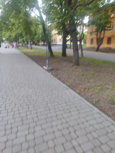 The image shows a paved pedestrian walkway with a grassy area and trees to the right. In the background, there are apartment buildings, likely residential buildings typical of Zhovti Vody, Ukraine. There are a few indistinct figures on the walkway, including what appears to be a person in pink clothing and another figure pushing a stroller. The lighting suggests it is daytime, possibly late afternoon or early evening, given the soft light and shadows. The perspective is low, looking forward along the walkway. The overall scene depicts a common urban environment in a Ukrainian city, characterized by residential buildings, green spaces, and pedestrian infrastructure.