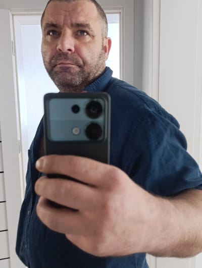 In a self-portrait taken from a medium close-up perspective, a man with short brown hair and a stubbled beard is seen holding a smartphone. He is wearing a dark blue collared shirt. The background suggests an indoor setting with a white door frame visible on the left. The image is cropped to focus on the man's face and the phone he is holding, obscuring much of the environment. The location in Marshyntsi, Ukraine is not discernible from the visual information provided in the image.
