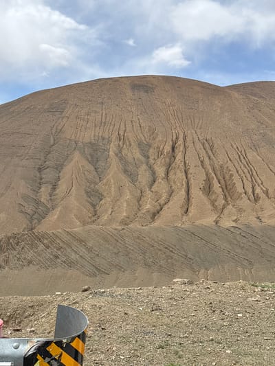 The image depicts a large mound of light brown earth or sediment, displaying significant erosion patterns with numerous rivulets and stratified layers. To the left of the mound is a portion of a blue sky with scattered white clouds. To the right, adjacent to the mound, is ground covered in loose stones and gravel. A roadside post, featuring black and yellow diagonal stripes, is partially visible at the lower right corner. The location is Tashi Dzom town, China, based on the query context.
