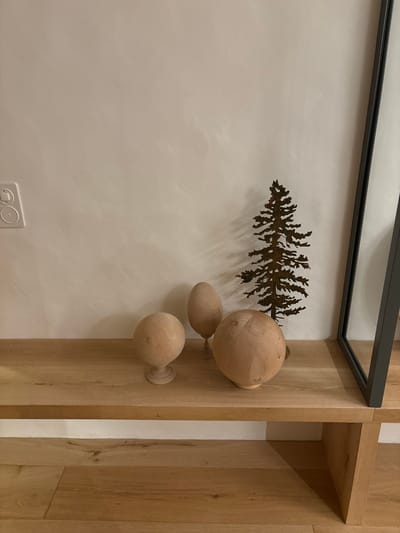 The image shows a wooden shelf with three wooden spheres and a metal silhouette of a pine tree on top. The shelf is a light wood color and is mounted on a wall. The wall is painted a light beige color and has a white electrical outlet on the left side. The shelf is part of a larger wooden structure. The wooden spheres are all different sizes and shapes, and the silhouette of the pine tree is in the center of the shelf. The image is taken from a low angle, looking up at the shelf. It is located in a building in the city of Celerina/Schlarigna, Switzerland, at coordinates 46.514, 9.859.