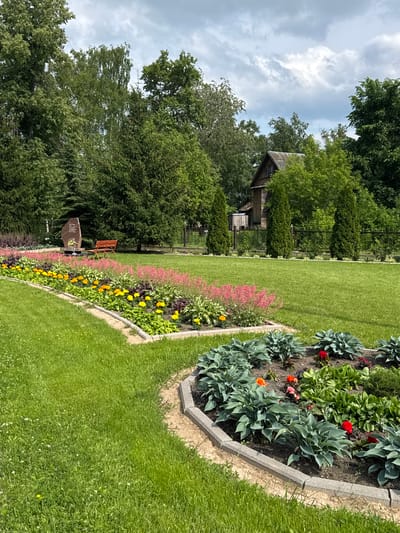 This is a full, eye-level shot of a landscaped park area with a large flowerbed in the foreground, a lawn, and trees in the background. The flowerbed contains various plants, including hostas, marigolds, and other flowering plants in shades of pink, yellow, and red. The lawn is neatly mowed and appears to be well-maintained. In the background, there is a wooden building with a rustic appearance, a fence, and several tall evergreen trees. The sky is partly cloudy. This scene is located in the city of Chudovo, Russia, and appears to be a peaceful and well-kept public space, possibly a memorial park due to the presence of a large stone monument and a bench. There are no people in the image.
