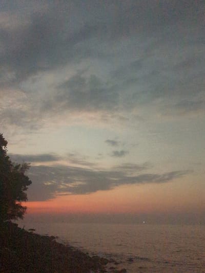 The image displays a coastal landscape at twilight, depicting a scene in a coastal area of Preah Sihanouk, Cambodia. The upper two-thirds of the frame are occupied by the sky, transitioning from dark grey-blue at the top to lighter grey and then to a band of orange and red hues along the horizon. Scattered grey clouds are visible across the sky. The lower right portion of the image shows a calm body of water, dark in tone but reflecting the ambient light. A distinct bright white point light source is visible on the water's surface towards the center-right, with fainter, smaller lights discernible further to the right. The lower left quadrant presents a dark landmass or rocky shoreline, characterized by irregular dark shapes at the water's edge. Dark, dense foliage, consistent with trees or bushes, is silhouetted against the sky along the upper edge of this landmass. No individuals or specific human activities are observable in the scene.