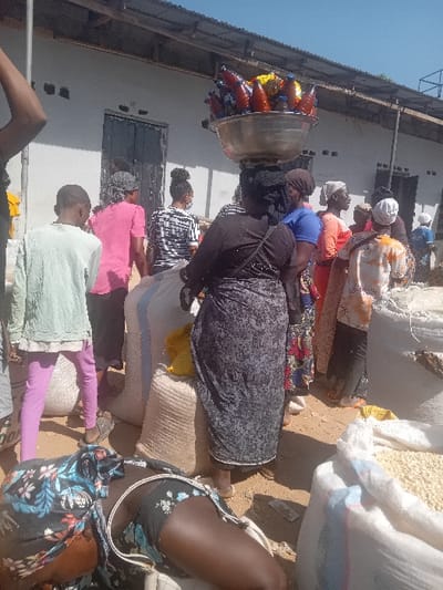 The image depicts an outdoor market scene in Gurku Gwari, Nigeria, under direct sunlight. Approximately 10-15 individuals are present, primarily adults. In the immediate foreground, a person with dark skin, wearing a dark floral top and dark shorts, is lying on their left side on the dusty, unpaved ground, with a white strap from a bag visible across their torso. In the central midground, a woman with dark skin carries a large, reflective silver basin on her head, filled with approximately 15-20 plastic bottles containing various colored liquids (red, orange, yellow, and blue). She wears a dark top and a patterned dark skirt, facing away from the viewer. To her left, a young individual in a light green long-sleeved shirt and purple pants stands facing away. Numerous large white woven sacks, consistent with those used for bulk goods like grains or legumes, are stacked throughout the scene; one in the foreground contains pale yellow granular material. Other individuals are distributed across the midground and background, dressed in various casual and traditional garments, including head wraps, patterned skirts, and t-shirts. A light-colored, single-story building with a corrugated metal roof and multiple window/ventilation openings, along with a dark door, forms the background. The environment is unpaved and dusty, consistent with an active outdoor market. Strong shadows are cast due to bright illumination.