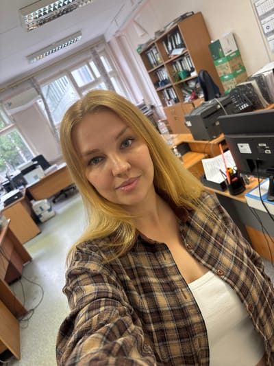 A young woman with blonde hair and blue eyes takes a selfie in an office environment. She is wearing a brown and tan plaid shirt over a white ribbed tank top. The office appears to be a standard workspace with desks, computers, filing cabinets, and fluorescent lighting. Through the window, trees and buildings are visible, suggesting an urban or suburban setting. The overall impression is that of a typical workday.