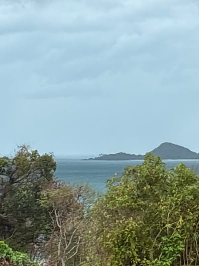 The image presents a coastal scene, likely taken from an elevated position. The sky is overcast, suggesting a cloudy or rainy day. A body of water, presumably the ocean or a bay, is visible with a landmass or island appearing in the distance. Trees and foliage are prominent in the foreground, partially obscuring the view. The overall impression is one of a natural setting, potentially a park or coastal area in Ban Bang Khao, Thailand. The absence of clear landmarks or human activity makes pinpointing the exact location challenging.