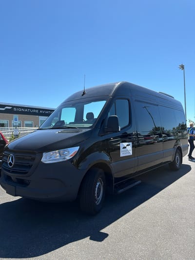 In Town of Southampton, United States of America, a black Mercedes-Benz Sprinter van is parked in an asphalt lot under a clear blue sky. The van has tinted windows and a "SIGNATURE AVIATION" logo on its side. In the background, a building with the same "SIGNATURE AVIATION" signage is visible, along with parking signs indicating reserved spots for disabled parking. A person in a safety vest and dark pants is walking away from the van towards the right side of the frame. The scene is illuminated by bright sunlight, casting a shadow from the van onto the pavement.