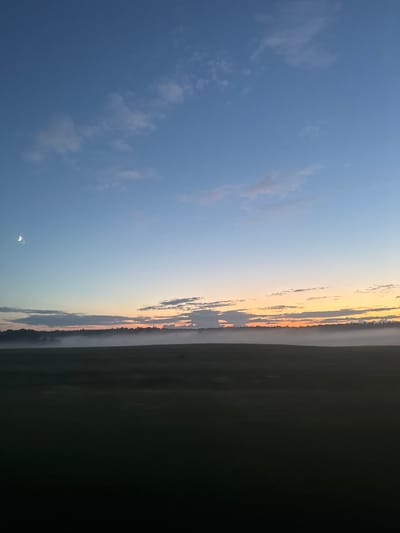 This is a wide shot of a field at dusk. The sky transitions from deep blue at the top to a pale orange near the horizon, with scattered clouds. A sliver of the moon is visible in the upper left corner. A thin layer of fog hovers over the dark, indistinct landscape of fields and trees that forms the lower half of the image. There are no people or animals visible in the scene. The location is identified as Егорье, Russia.