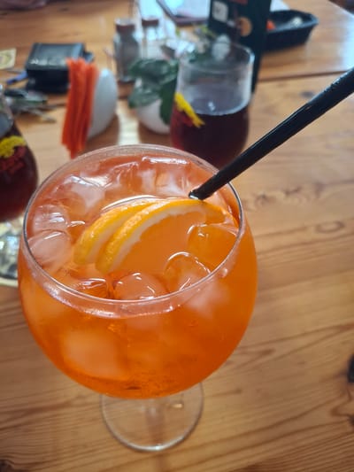 The image shows a close-up of an orange cocktail in a large wine glass, filled with ice cubes and garnished with three thin orange slices. A black straw extends from the drink. The glass rests on a wooden table, with the background blurred. To the left of the cocktail, partially visible, is another glass with a dark red liquid and a yellow garnish. Further in the background, also blurred, are salt and pepper shakers, a small potted plant, and a black object that could be a phone or wallet. The overall scene suggests an outdoor or casual indoor setting, possibly a cafe or restaurant, with a focus on the refreshing drink. There is no identifiable information about the location within the city of Lēdmane, Latvia.
