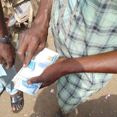 This image captures an outdoor scene in Olomoda, Nigeria, depicting a financial interaction between two individuals in daylight. The central figure, with dark skin, is prominently visible from the mid-torso downwards, wearing a light green and dark plaid patterned long-sleeved garment or wrapped fabric. Their hands are actively handling a stack of Nigerian 1000 Naira banknotes, indicating counting or an exchange. The banknotes clearly display "CENTRAL BANK" and the "1000" denomination.
To the left, a second individual, also with dark skin, is partially visible. Their left arm, adorned with a black wristwatch, and their left foot, wearing a black flip-flop branded "Dior," are discernible. The ground beneath the individuals appears to be an unpaved or dirt surface, with scattered refuse, including crumpled papers and small pieces of debris. In the upper left background, indistinct discarded materials, possibly remnants of market stalls or construction debris, are present.