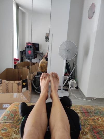The image depicts a personal living space, likely a bedroom, in Grosseto, Italy. In the foreground, a person's legs and feet are visible, resting on what appears to be a patterned comforter. The person is positioned with their feet slightly elevated, suggesting they are lying down or sitting in a reclined position.

The background reveals a room that seems to be in a state of transition or disarray, with several large cardboard boxes stacked. A black gaming chair with red accents is visible, along with a computer monitor and desk in a mirrored reflection, indicating the presence of a workspace. A floor fan stands to the right of the frame, and a small white space heater is positioned near its base.

The overall impression is of a lived-in environment, potentially a room undergoing packing or unpacking, with everyday objects and furniture filling the space. The lighting appears to be natural, coming from a window or overhead source, creating subtle shadows and highlights on the objects in the room. The presence of a vacuum cleaner and other cleaning supplies suggests ongoing household activity.
