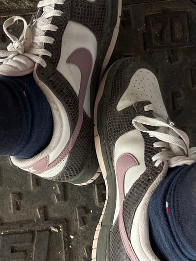 A top-down view shows the feet of an individual wearing dark grey and white athletic shoes with a snakeskin-like texture, white laces, and a pink Nike "Swoosh" logo. The shoes are positioned on a dark, metallic manhole cover or utility grate. The right shoe is worn over a dark blue sock displaying a red, white, and blue striped pattern near the ankle. The left shoe also covers a dark blue sock. The metallic surface features raised alphanumeric text, clearly identifying the location as "FOLIGNO" and "70," indicating the city of Foligno, Italy. Scattered white debris is present on the dark surface. The individual's posture suggests standing.