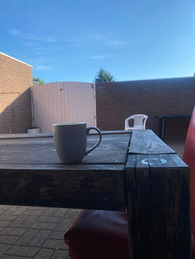 The image depicts a mug of coffee on a rustic wooden table outdoors. In the background, a light-colored double gate is visible, flanked by a brick building on the left and a brick wall on the right. A white plastic chair sits in front of the brick wall, with a dark table and a red chair partially visible to the right of it. The scene is set in a backyard with paved ground. The sky is blue with sparse clouds, indicating daylight. The location is identified as the city of Venlo, Netherlands.