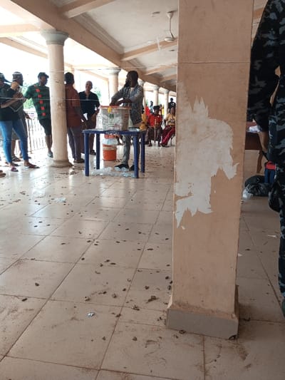 The scene unfolds in a public or communal covered space within the city of Ajalli, Nigeria, identifiable by its tiled floor, multiple light-colored support columns, and a visible ceiling fan. At the center, a bearded male, dressed in a denim-style shirt, stands behind a blue plastic table, actively handling a transparent plastic box labeled with white paper, consistent with a ballot or collection box. Two white buckets are stacked beneath the table.
Approximately 10-15 other individuals are present. To the left, a group of five adults stands near a column, observing the central figure's activity. In the background to the right, several individuals, primarily women, are seated on red chairs. The tiled floor is extensively scattered with numerous small, dark, oblong objects and several discarded clear plastic water bottles. A large, light-brown rectangular pillar, exhibiting areas of peeling paint or removed adhered material, dominates the right foreground, partially obscuring a person in camouflage attire.