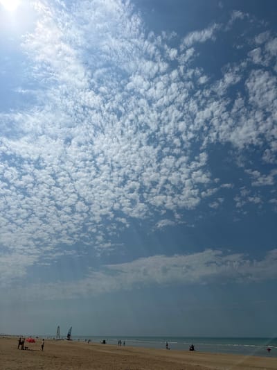 This is a photograph of a beach scene in Koksijde, Belgium, under a partly cloudy sky. The sky is predominantly blue with scattered cumulus clouds. Sunlight is visible, casting rays through the atmosphere. The beach itself is sandy and appears to be a popular spot, with numerous people present. There are several sailboats visible near the water's edge, suggesting recreational activities are taking place. Some individuals are walking along the shore, while others are gathered in groups. A red umbrella provides shade for a small group on the beach. The sea is calm, with gentle waves lapping the shore. The overall impression is a pleasant day at the beach.