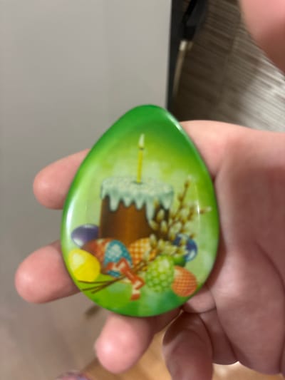 A person's hand is holding a green, egg-shaped Easter-themed magnet. The magnet features an illustration of a traditional Easter cake with white frosting and a lit candle, surrounded by decorated Easter eggs, pussy willow branches, and a bright green background. The image is slightly blurred.