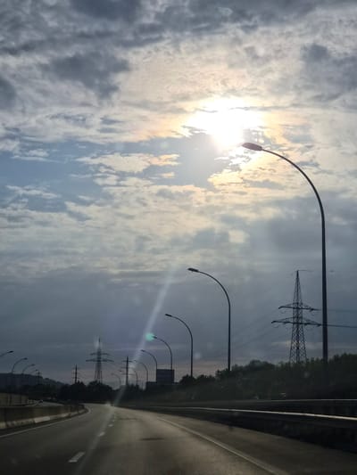The image depicts a view from a vehicle on a highway in Dudelange, Luxembourg. The sky is overcast with the sun breaking through the clouds, creating a bright, hazy glow. Several tall, curved streetlights line the highway. In the background, large electricity pylons with power lines are visible, set against a backdrop of trees and a building. The highway itself is a multi-lane road with concrete barriers separating the lanes. The overall scene suggests a late afternoon or early evening with the sun low on the horizon.