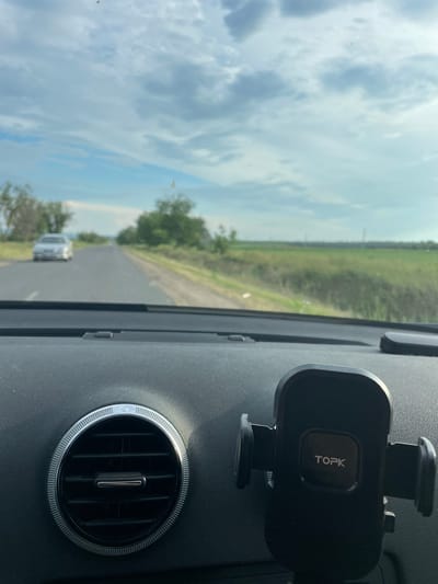 The media file is a first-person perspective shot from inside a vehicle. The camera is angled slightly to the right, capturing the dashboard and a view through the windshield. On the dashboard, a black phone mount with the "TOPK" logo is visible on the right side. To the left of the phone mount, a circular air vent with a chrome trim is prominent.

Through the windshield, a two-lane road stretches into the distance, flanked by fields of green grass and scattered trees. The sky is partly cloudy, with patches of blue visible between the white and gray clouds. The scene suggests an overcast day with some sunshine. A silver car is driving on the road ahead, creating a sense of movement and travel. The overall setting appears to be a rural or semi-rural area, possibly in Moldova given the location context. The time of day is difficult to determine precisely but likely daytime due to the lighting conditions. There are no people visible in the frame. The image conveys a sense of a journey in progress.