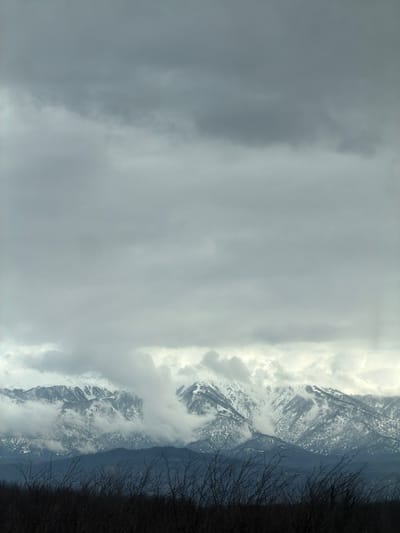 The scene depicts a mountain landscape under an overcast sky, viewed from the vicinity of Samtredia, Georgia. The upper two-thirds of the image are dominated by a dense, gray cloud cover, with variations in tone suggesting multiple cloud layers. In the mid-ground, a prominent range of mountains is visible, heavily covered in white snow, particularly on their peaks and upper slopes. Wisps of white and gray clouds are intertwined with the mountain ridges, partially obscuring them and indicating low-lying fog or cloud banks. Below the snowline, darker patches of terrain, possibly forested areas, are visible. A darker, bluish-gray mountain range or foothills lies in the lower mid-ground, receding into the distance. The immediate foreground features a dark, indistinct band of bare, tangled branches or shrubbery, silhouetted against the lighter background. No individuals, human activity, or man-made structures are discernible in the scene.